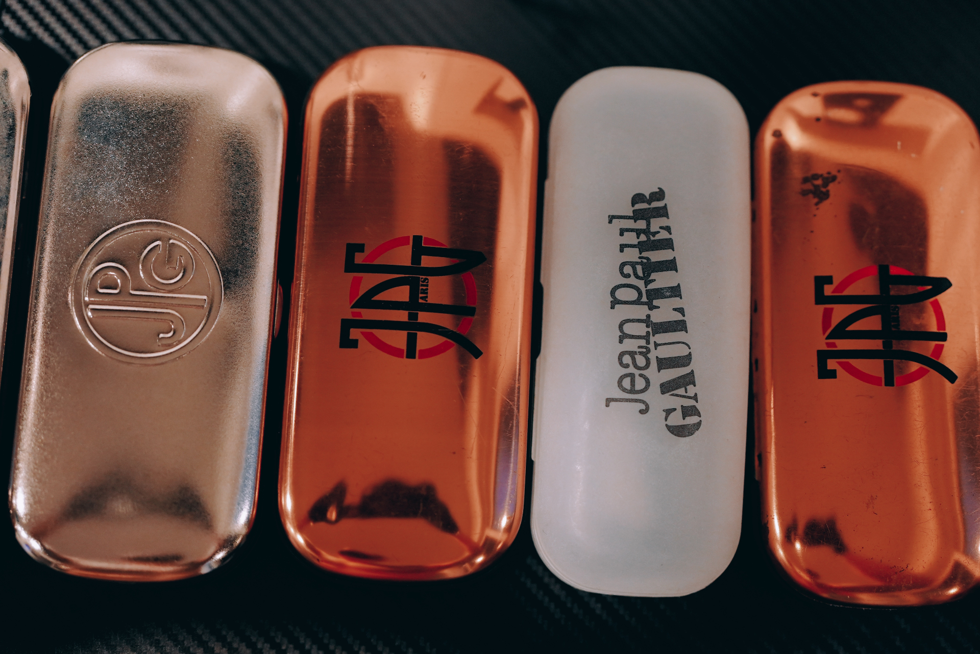 Five vintage Jean Paul Gaultier eyewear cases lined up — silver JPG monogram, copper JPG Paris with red circle, and frosted Jean Paul Gaultier logo.