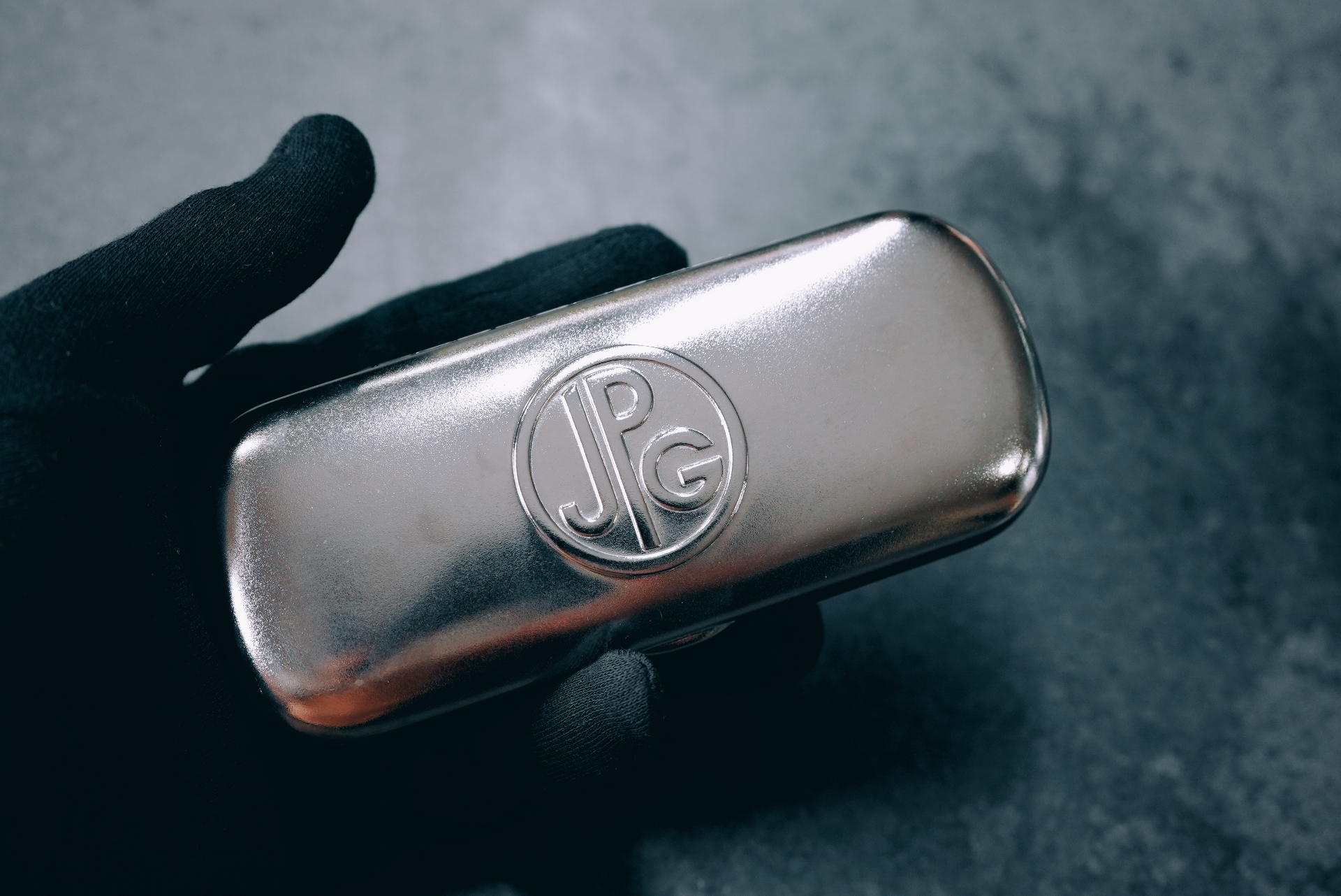 Close-up of a silver vintage Jean Paul Gaultier case with the JPG monogram seal, held in a gloved hand.